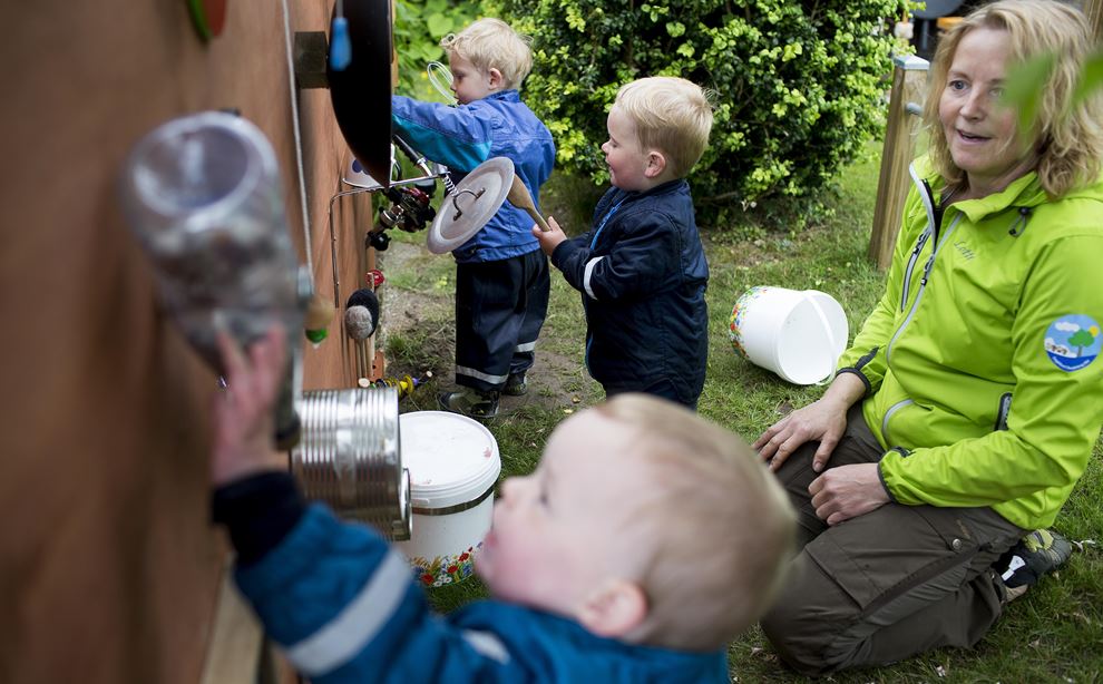 Dagplejer og tre drenge leger ved udendørs musikvæg lavet af gamle gryder og dåser. 